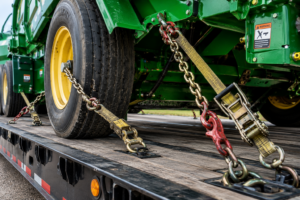 Close-up of DOT-compliant chain tie-downs securing a hay baler's frame to a flatbed trailer deck.