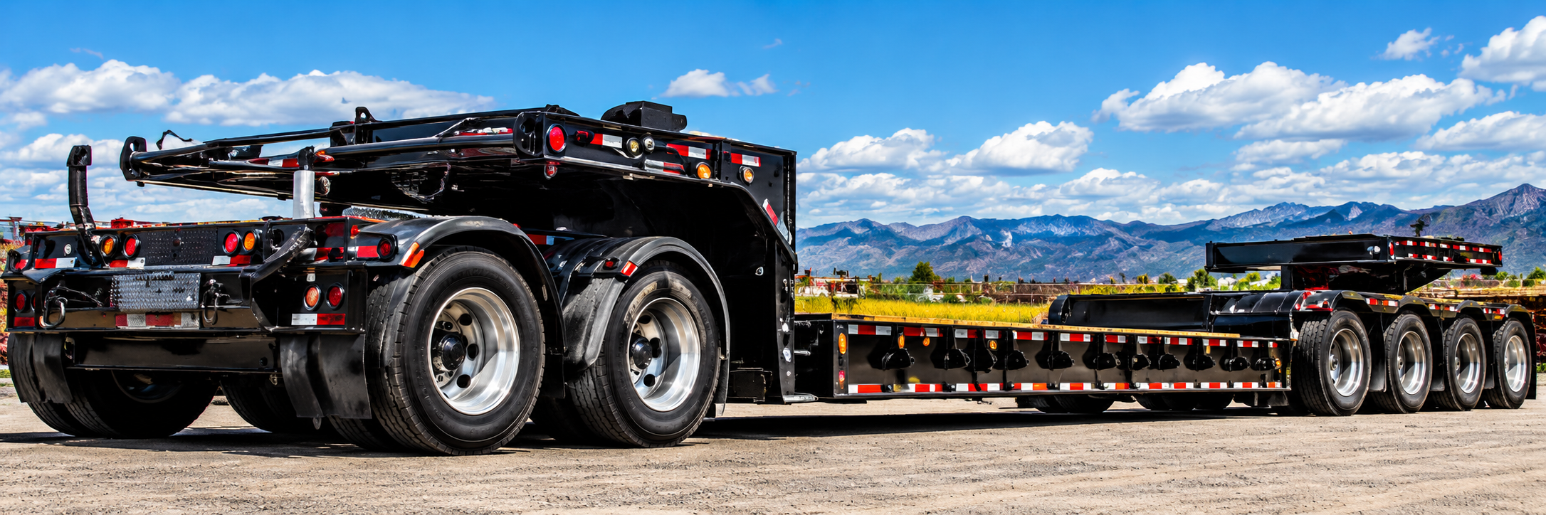 Empty black multi-axle lowboy trailer with a rear booster axle and wood deck parked on a gravel yard in front of Montana mountains, used for heavy haul trucking